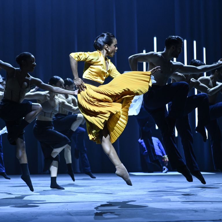 A woman with a yellow dress dancing in front of a group of male dancers. 