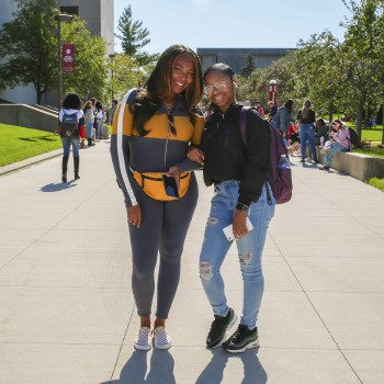 Two female students standing and smiling outdoors on a college campus. 