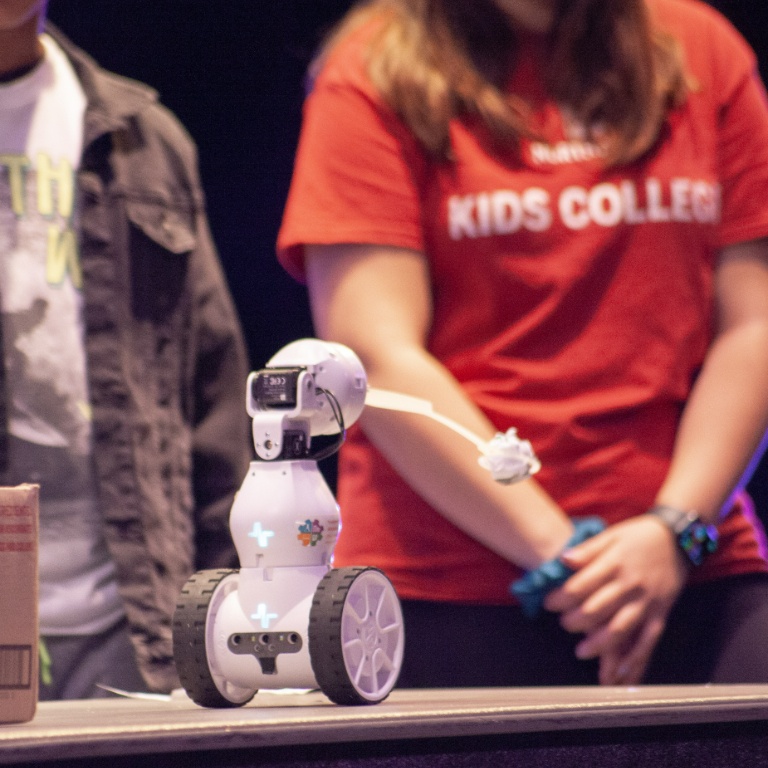 A robot with wheels moves across a desk with students in the background.