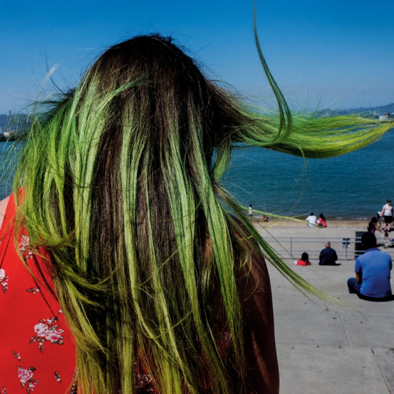 Photograph of a person with long green hair sitting in front of a body of water.